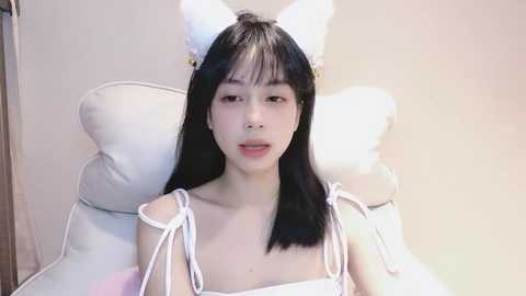 Media: Video of a young East Asian woman with straight black hair, wearing white cat ears and a matching sleeveless top, sitting on a white cushioned chair against a plain beige wall.