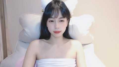 Media: A young Asian woman with long black hair and a slender physique sits on a white cushioned chair, wrapped in a white towel, against a beige wall.