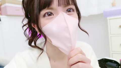 Media: Video of a young woman with fair skin, brown hair, and bangs, wearing a white lab coat and a pink surgical mask. She is indoors, with white walls and pastel furniture in the background.