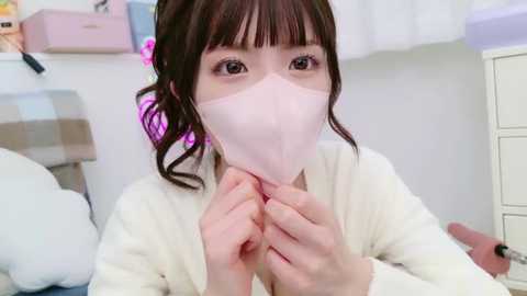 Media: Video of an Asian woman with dark hair in pigtails, wearing a pink mask and white robe, holding her mask, in a cluttered bedroom with pastel decor.