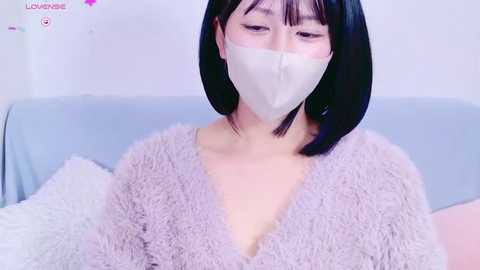 Media: A video of an East Asian woman with straight black hair, wearing a white face mask, a fuzzy lavender robe, and sitting on a light blue couch.