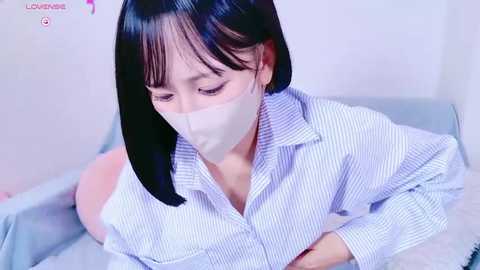 Media: A video of an East Asian woman with straight black hair, wearing a white striped button-down shirt and a face mask, sitting on a bed, looking down.