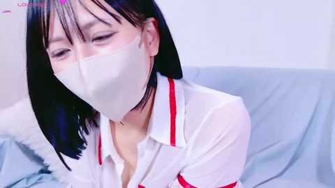 Media: Video of a young Asian woman with shoulder-length black hair, wearing a white nurse's uniform with red trim, and a surgical mask, sitting on a bed with light blue sheets.