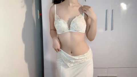 Media: Video of a slender Asian woman with fair skin, wearing white lace lingerie: a bra and panties, in a modern, minimalist room with white walls and cabinets.