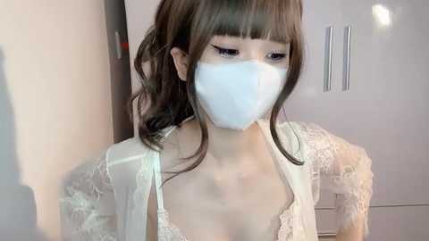 Media: Video of an East Asian woman with pale skin, brown hair, and bangs, wearing a white lace bralette and a face mask, standing indoors.