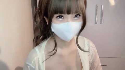 Media: Video of a young Asian woman with long brown hair, wearing a white face mask, light green robe, and a light green top, standing indoors with a light-colored wall and cabinet in the background.