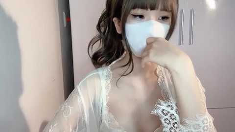 Media: Video of an Asian woman with long, dark hair, wearing a white lace robe and surgical mask, covering her nose and mouth, indoors.