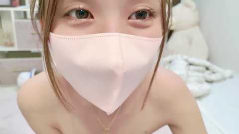 Media: Video of an Asian woman with fair skin and brown hair, wearing a pink face mask, looking directly at the camera, in a softly lit bedroom with white bedding and a headboard.