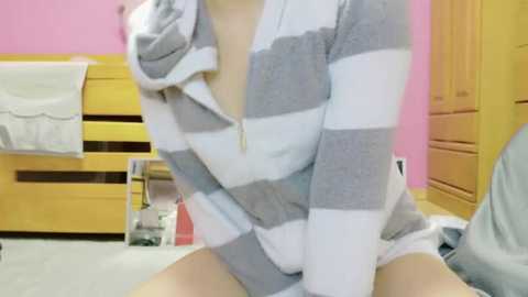 Media: Video of a light-skinned woman with small breasts, wearing a grey and white striped hoodie, revealing her cleavage, in a pink and yellow room with wooden furniture and a white towel hanging on a drawer.