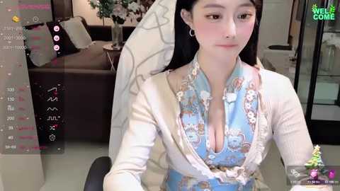 Media: Video of a young Asian woman with long black hair, fair skin, and wearing a low-cut, floral-patterned blue dress. She sits in a modern living room with white furniture and plants.