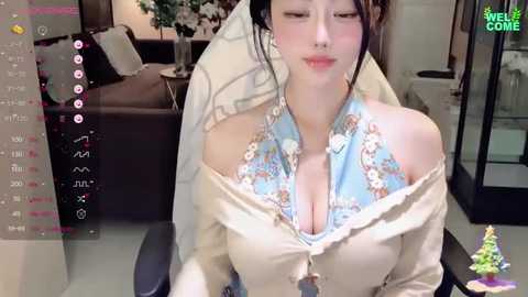 Media: Video of a young Asian woman with fair skin and black hair in a floral off-shoulder dress, sitting in a modern living room with a TV screen showing a countdown.