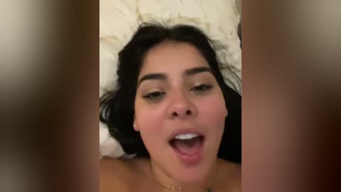 Media: Video of a Latina woman with olive skin, dark hair, and full lips, lying on a beige pillow, partially obscured by out-of-focus, tan hands.