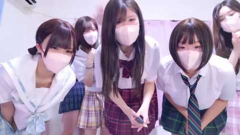 Media: Video of five Japanese schoolgirls in white face masks, red plaid skirts, and white shirts, standing in a row, indoors, with white walls and a pink shower curtain in the background.