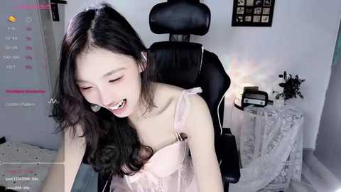 Media: Video of an Asian woman with long black hair, wearing a pink lace lingerie top, leaning forward in a black office chair, in a bright, minimalist room with white walls.