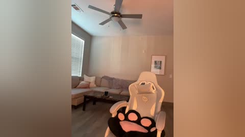 Media: Video of a modern living room with beige walls, a white gaming chair, and a gray sectional sofa with pillows, a large window with blinds, and a ceiling fan.