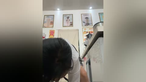 Media: A video of a person with dark hair, wearing a white shirt, leaning over a kitchen counter, with a cluttered background featuring framed posters and kitchen items.