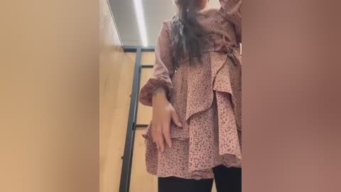 Media: Video of a woman with long, dark hair, wearing a pink floral-patterned, long-sleeved dress with ruffled hem, black leggings, and holding her dress's hem in a narrow hallway with beige walls and a black-framed window.