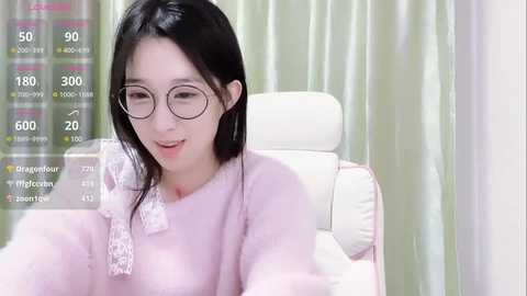 Media: Video of a young East Asian woman with straight black hair, fair skin, and round glasses, wearing a light pink, fluffy robe, seated in a white chair against a green curtain backdrop.