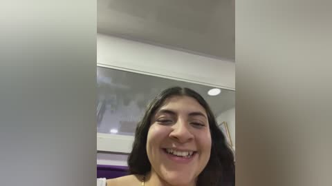 Media: Video of a young woman with long dark hair and a light complexion, smiling widely, taken indoors against a white wall and a glass door.