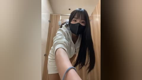 Media: Video of an Asian woman with long black hair, wearing a black face mask and a white hoodie, leaning into a stall with a wooden door and beige walls.