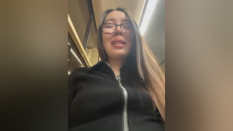 Media: Video of a young Asian woman with long, straight brown hair and glasses, wearing a black ribbed sweater. She stands in a beige, dimly lit subway car with fluorescent lights overhead.