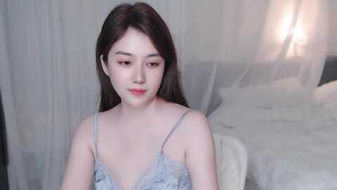 Media: Video of a young Asian woman with long, straight black hair, fair skin, and delicate facial features, wearing a light blue lace camisole, sitting on a bed with white bedding and sheer curtains in the background.