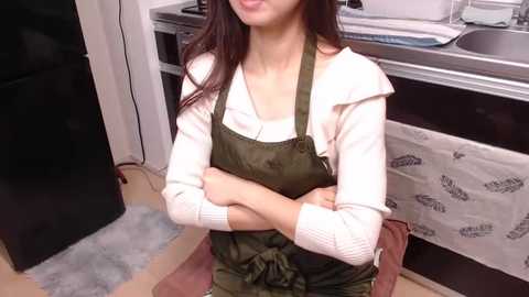 Media: Video of a young Asian woman with long dark hair, wearing a white sweater and olive green overalls, arms crossed, in a modern kitchen with stainless steel appliances and a white towel with text.