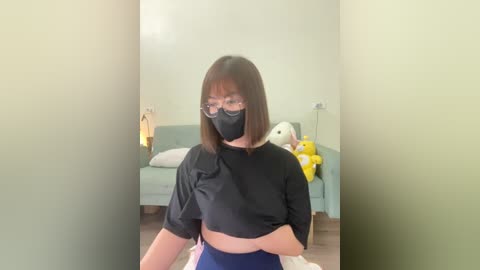 Media: Video of an Asian woman with straight, shoulder-length brown hair, wearing glasses, a black face mask, and a black cropped top, standing in a minimalist living room with a pastel green sofa, stuffed animals, and a soft, natural light.