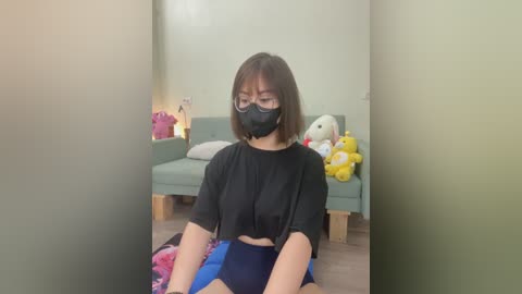 Media: Video of an Asian woman with short brown hair, wearing glasses, black face mask, and black top, seated on a plush blue cushion in a brightly lit, minimalist room with stuffed animals and a grey couch.