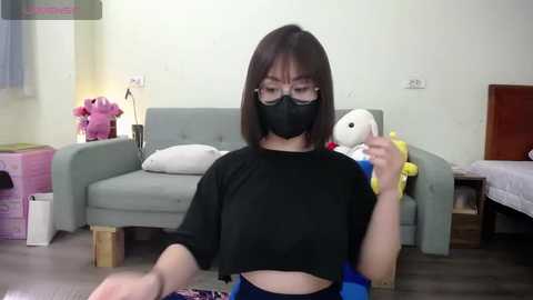 Media: Video of a young Asian woman with straight brown hair, wearing a black mask and short-sleeved top, sitting on a gray sofa in a brightly lit, minimalist room.