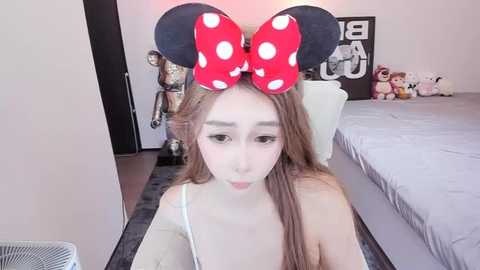 Media: Video of a young East Asian woman with fair skin and long brown hair, wearing a large red Minnie Mouse headband and a white tank top, sitting on a bed in a modern bedroom with gray bedding and stuffed toys.