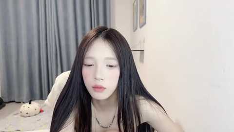 Media: Video of a young East Asian woman with long black hair, pale skin, and full lips, leaning against a white wall in a dimly lit room with gray curtains and a stuffed animal.