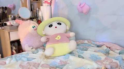 Media: Video of a pink teddy bear with a yellow bib and a yellow duck plush toy, both on a bed with a pastel-patterned blanket. Background includes a vanity with makeup items, a pink cat plush, and a large mirror.