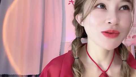 Video of a fair-skinned, young woman with blonde pigtails, wearing a red top and necklace, set against a soft, pink-tinted background.