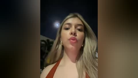 Media: Video of a blonde woman with fair skin, wearing a red top, blowing a kiss towards the camera under a night sky.