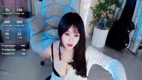 Media: Video of an Asian woman with long black hair and fair skin, wearing a low-cut white dress, kneeling in a modern room with a white chair, guitar, and plants.