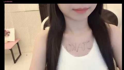 Media: A video of an East Asian woman with long black hair, fair skin, and a white tank top, showing a tattoo of the word \"LOVE\" on her chest. She is seated indoors with a pink chair and a pink object in the background.