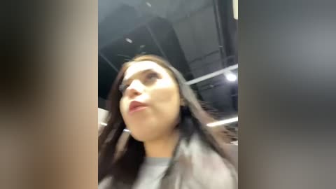 Media: A blurry video of a woman with medium-length brown hair and fair skin, wearing a grey jacket, taken from a low angle, likely inside a dimly lit room with metallic ceiling panels.