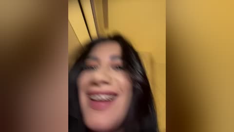 Media: Video of a young woman with long, dark hair, smiling widely, with braces. She is in a yellow room, looking upward. The image is slightly blurred.