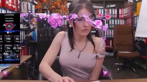 Media: Video of a Caucasian woman with long dark hair, wearing a sleeveless pink top, seated at a desk, surrounded by colorful flowers, books, and a computer screen displaying a game interface.