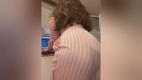 Media: Video of a woman with curly brown hair, wearing a pink-striped dress, bending over a blue countertop in a dimly lit room.