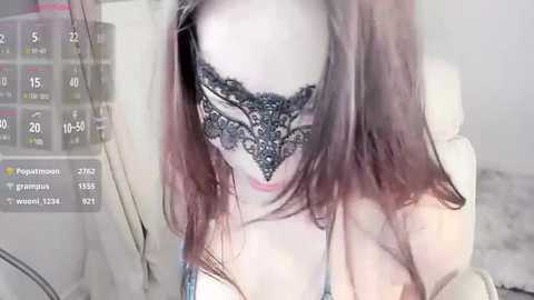 Media: A video shows a young woman with fair skin, wearing a black lace mask and a light blue tank top. Her long, straight, dark hair partially covers her face. The background features a white chair and a blurred medical device.