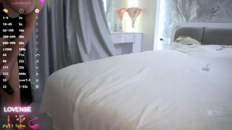 A video of a white, neatly made bed with a light gray headboard, partially covered by a white duvet, in a softly lit, modern bedroom with floral wallpaper. A person's hand is visible on the left side.