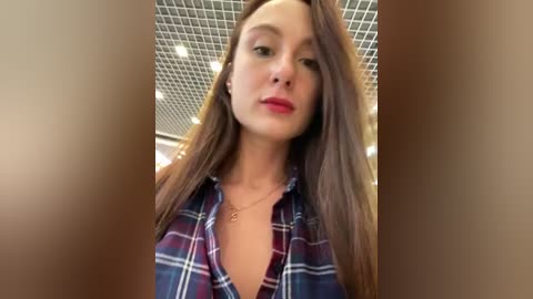 Media: A video of a young woman with long brown hair, wearing a plaid shirt and red lipstick, standing in a room with a tiled ceiling.