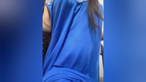 Media: Video of a person with long brown hair, wearing a loose-fitting, bright blue shirt, leaning forward, partially visible from the back. The background is blurred, with a blue gradient.