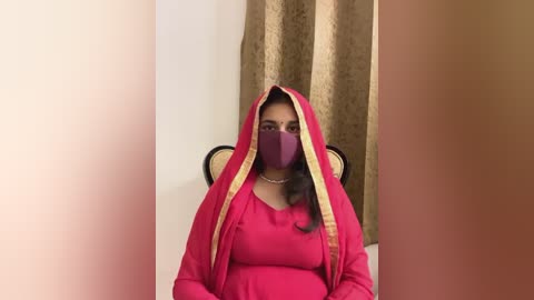 Media: Video of a woman wearing a red sari with gold trim, a purple face mask, and a pearl necklace, seated in a chair against beige curtains.