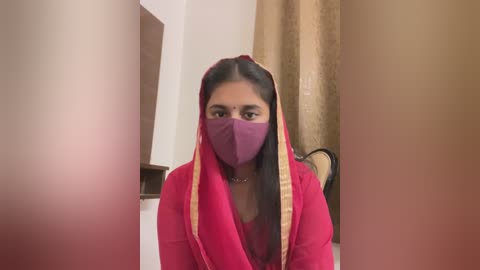 Media: Video of a young woman with long black hair, wearing a pink sari with a purple face mask, sitting in a beige room with a curtain.