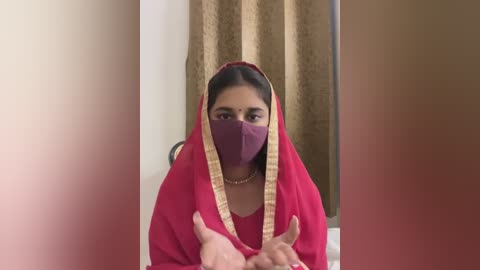 Media: A video of a South Asian woman wearing a maroon sari with gold border, a maroon mask, and a gold necklace, standing in a room with brown curtains.