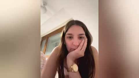 Media: A video of a young woman with long brown hair, resting her chin on her hand, wearing a gold watch, in a room with wooden doors and a fan.