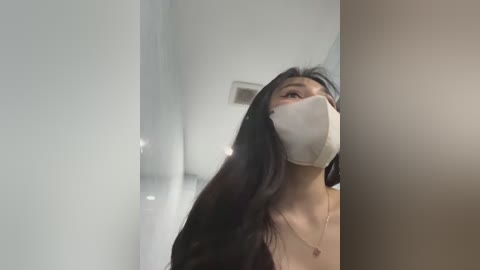 Media: A video of an Asian woman with long black hair and fair skin, wearing a white mask and necklace, gazing upward, standing beside a beige wall.